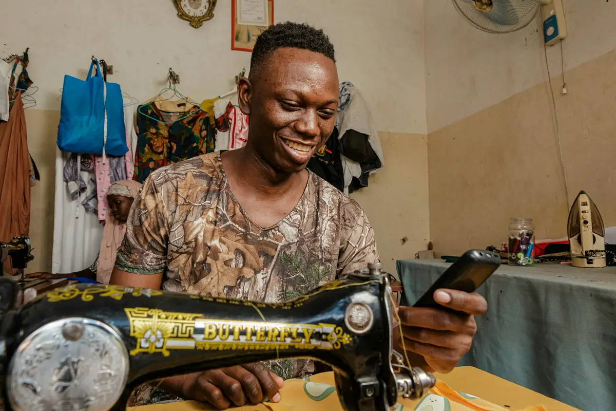 Un couturier sourit devant sa machine à coudre, son téléphone à la main